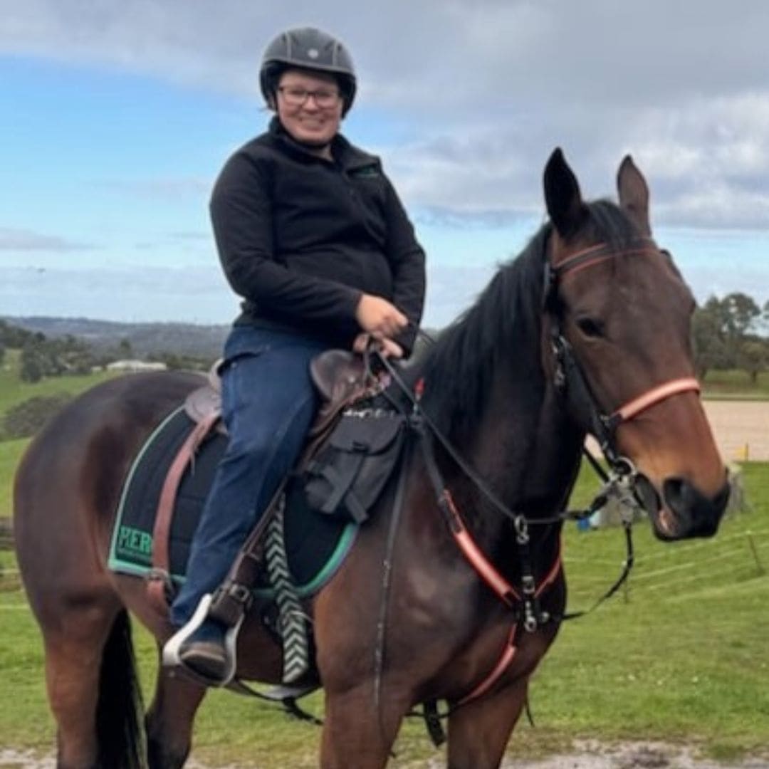 Woman wearing Ride Proud Riding Jeans riding a bay horse in an outdoor setting with green fields and trees.