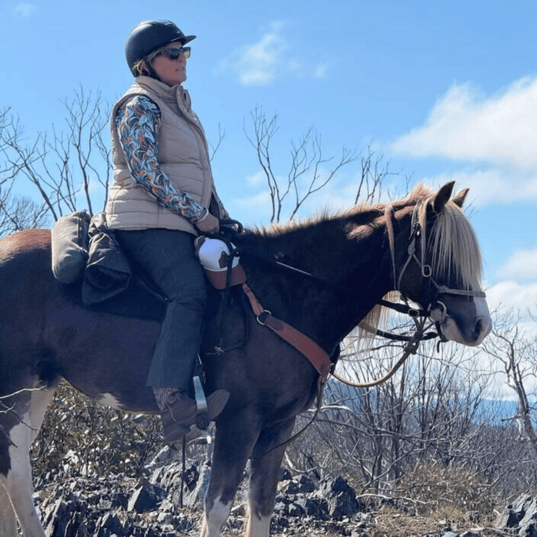 Person riding a horse in a mountainous area with a clear blue sky wearing riding shirt and pants from Ride Proud Clothing