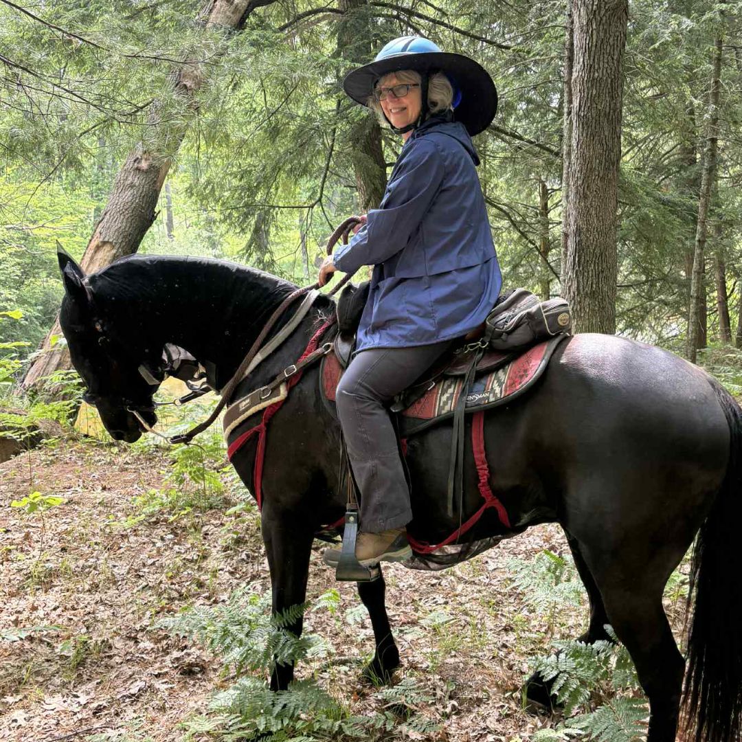Woman in Ride Proud Trainers riding pants sits confidently on her horse in forest, wearing blue jacket and wide-brimmed hat